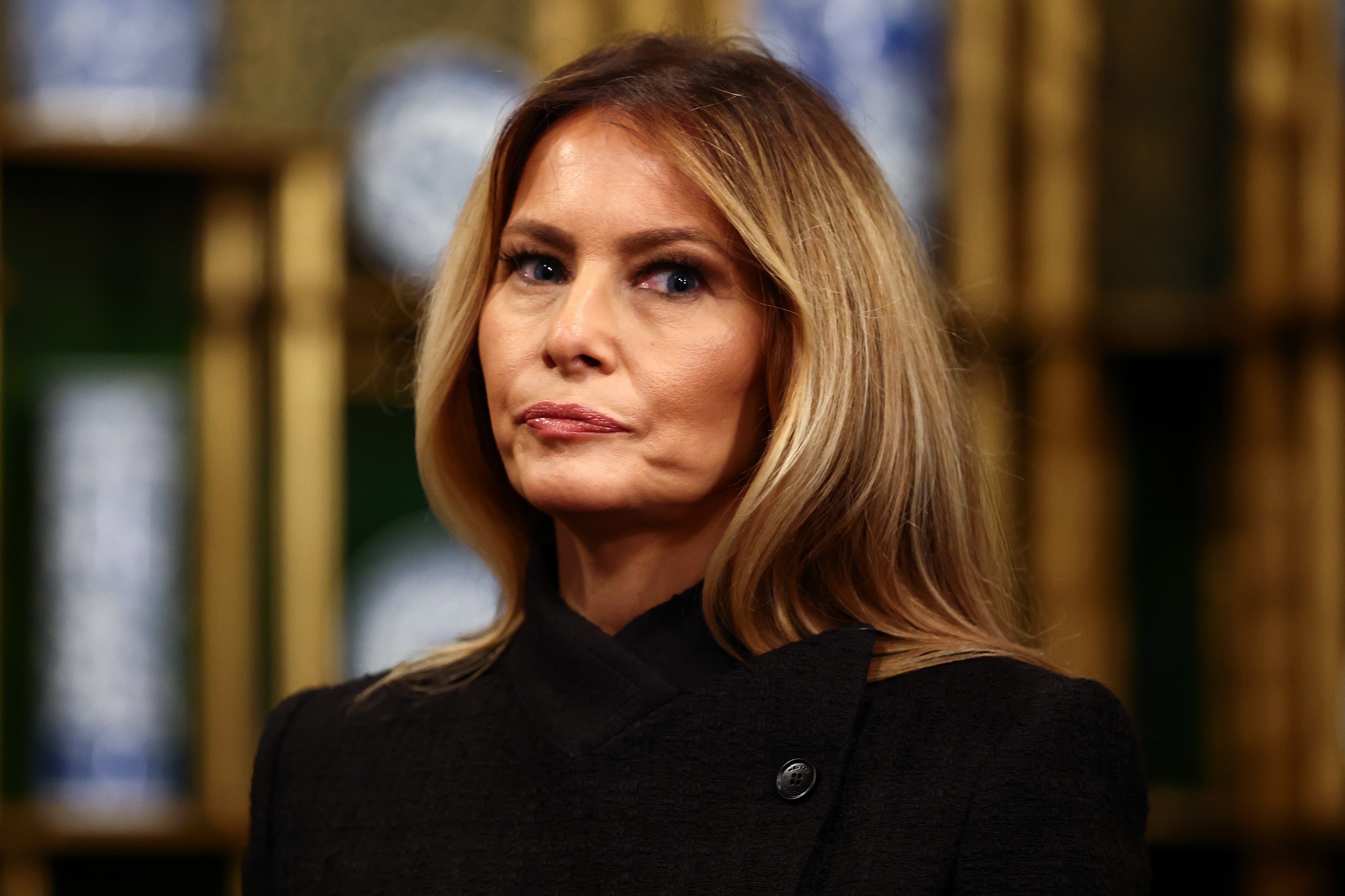 A person with long hair is wearing a dark, formal outfit, standing in front of a blurred background