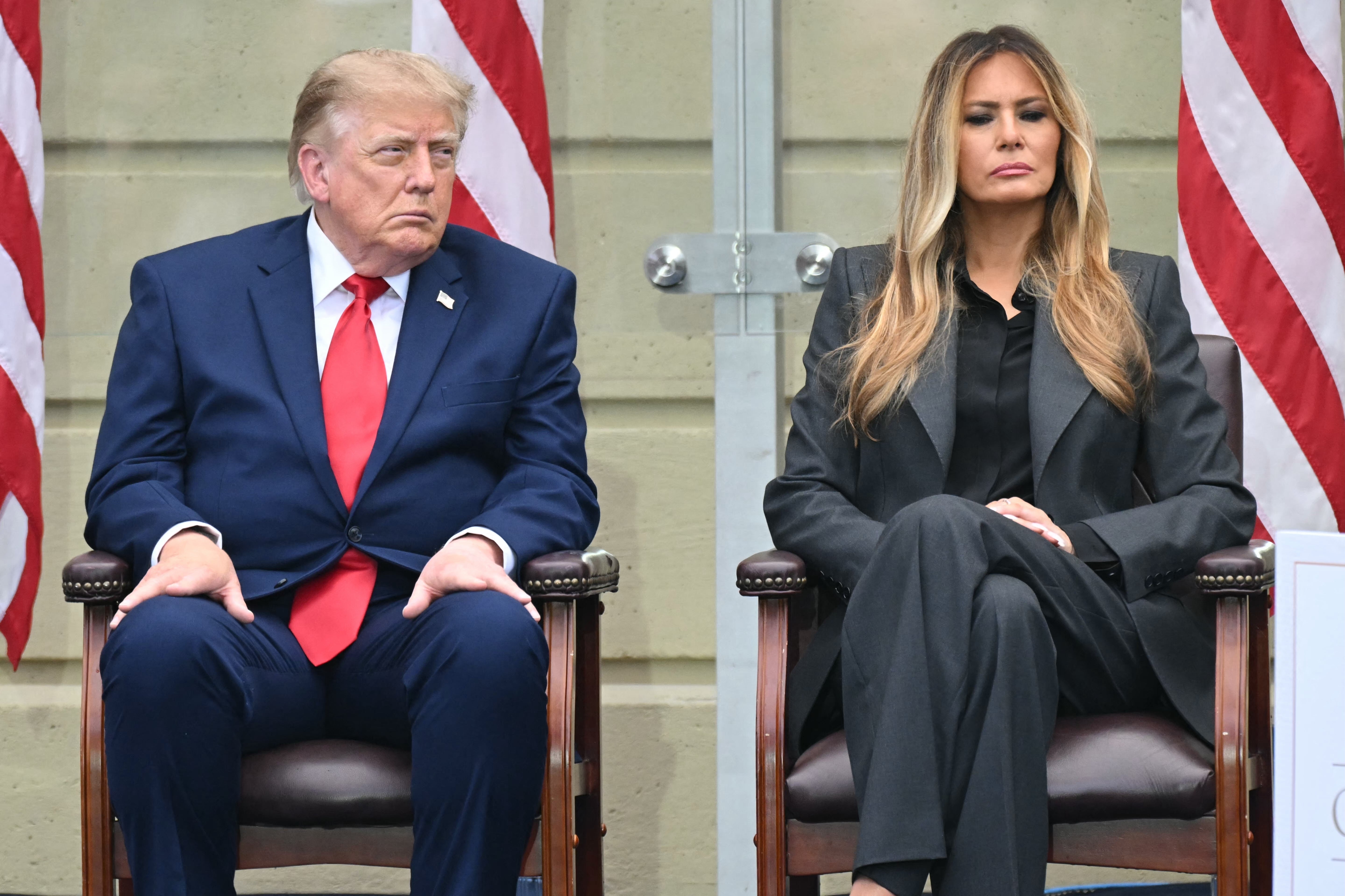 A man in a suit and tie sits next to a woman in a long-sleeved outfit, both on chairs, with flags in the background