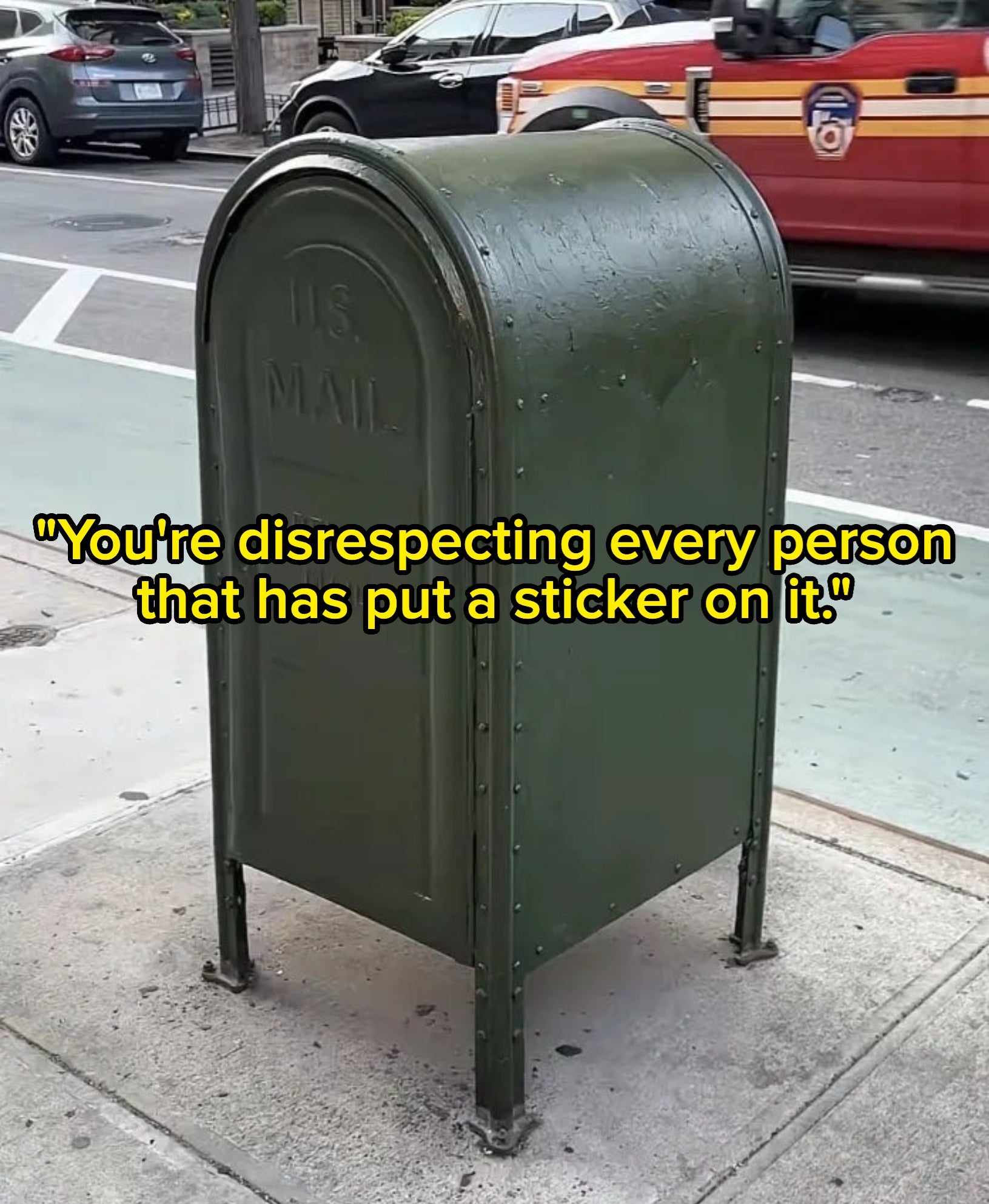A freshly-painted green U.S. mailbox is on a city sidewalk beside the street with cars and buildings in the background