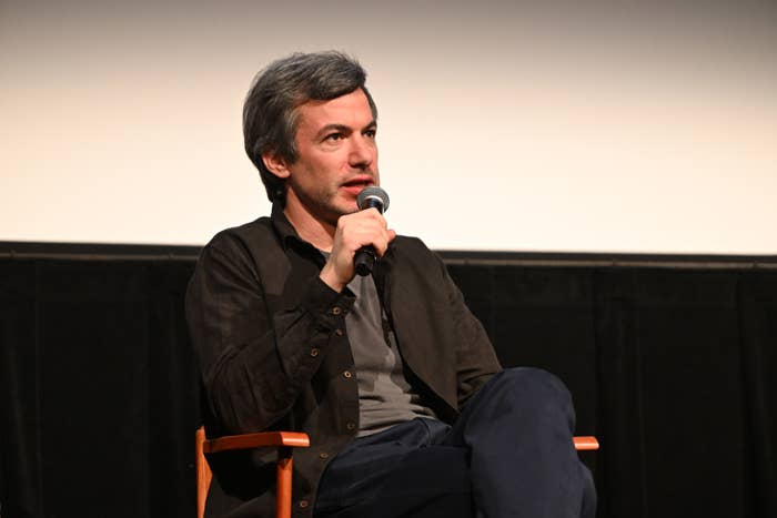 Nathan Fielder seated connected  stage, speaking into a microphone, wearing a acheronian  overgarment   and pants