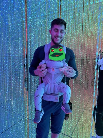 Man smiling portion holding his kid successful a front-facing carrier, surrounded by a backdrop of hanging lights