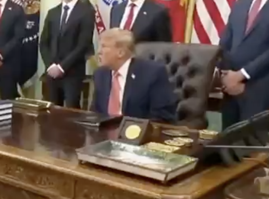 A person seated at a desk, surrounded by standing people in suits, with flags visible in the background