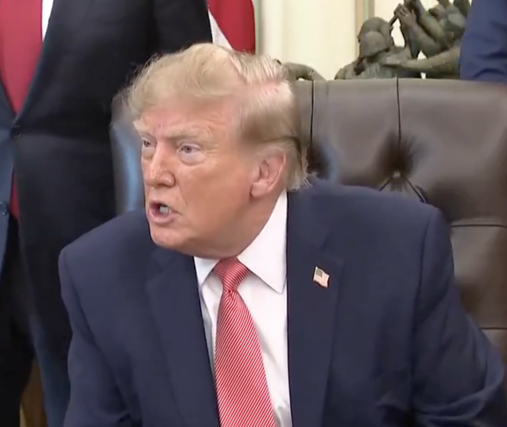 A person in a suit and red tie is sitting on a leather chair, appearing to speak during a formal setting