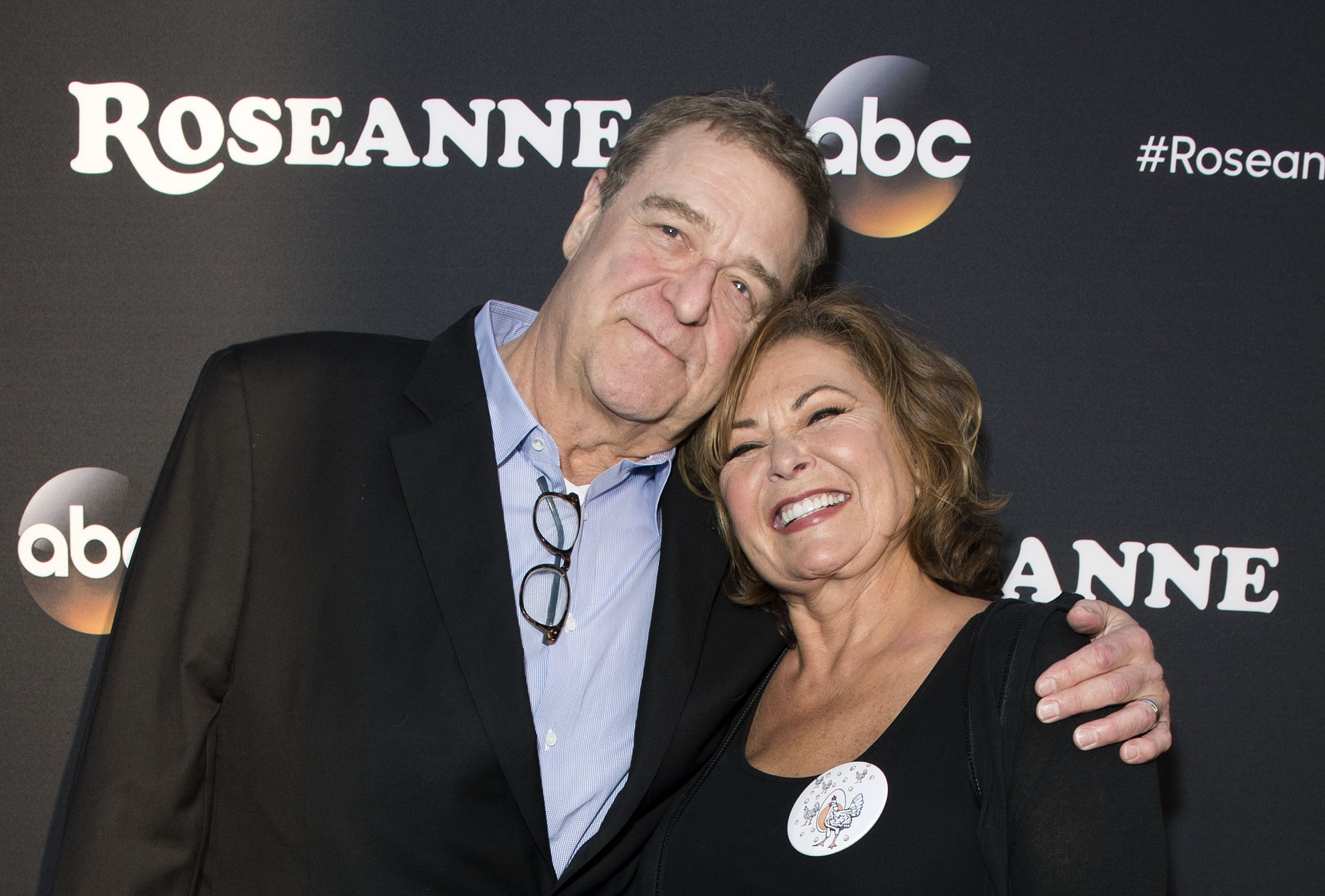 Actors John Goodman and Roseanne Barr smiling astatine a TV event, Goodman successful a acheronian suit with an unfastened collar and Barr with abbreviated hairsbreadth successful a blouse.