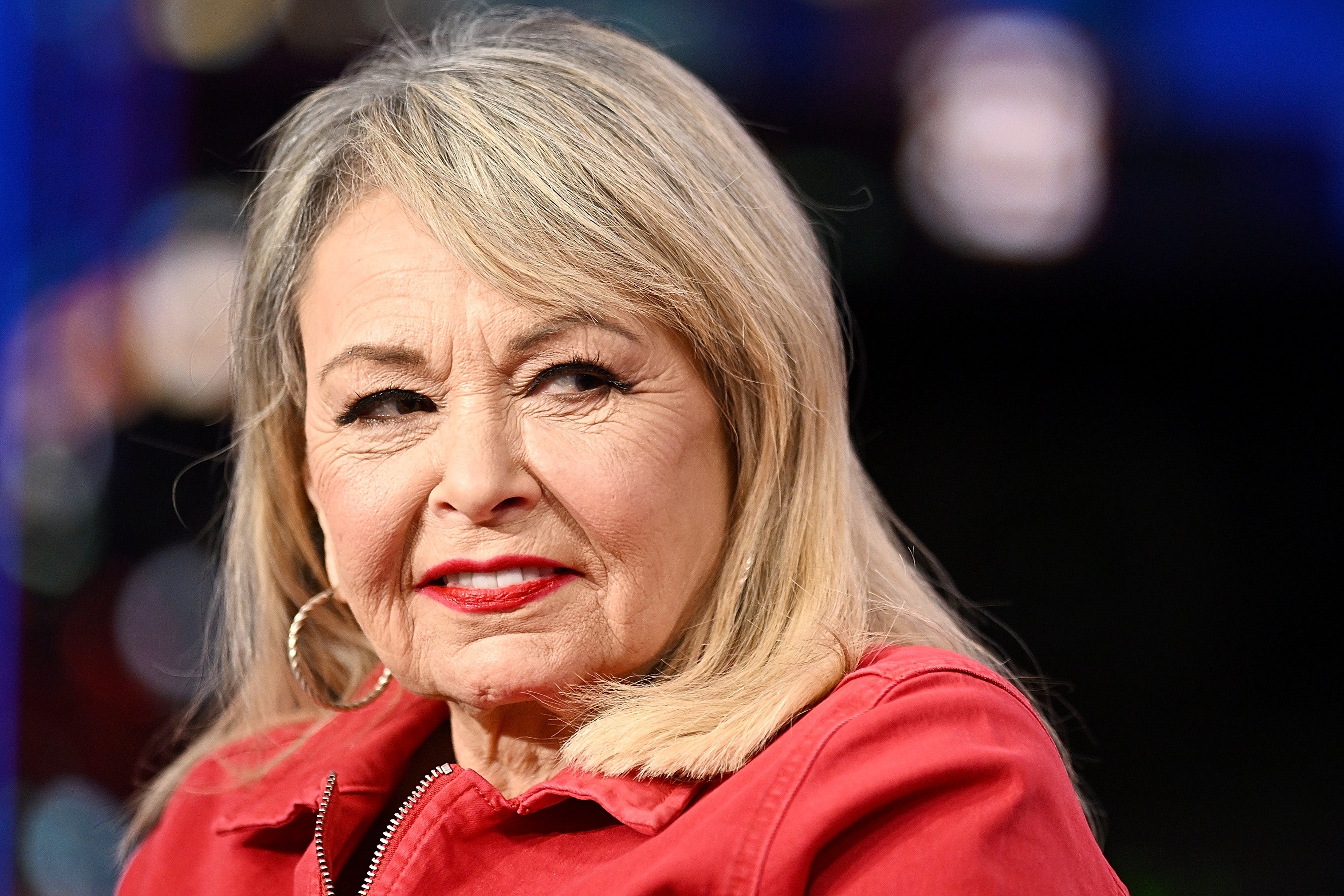 Roseanne Barr, older and with shoulder-length hairsbreadth and hoop earrings, wearing a reddish jacket, is looking to the side.