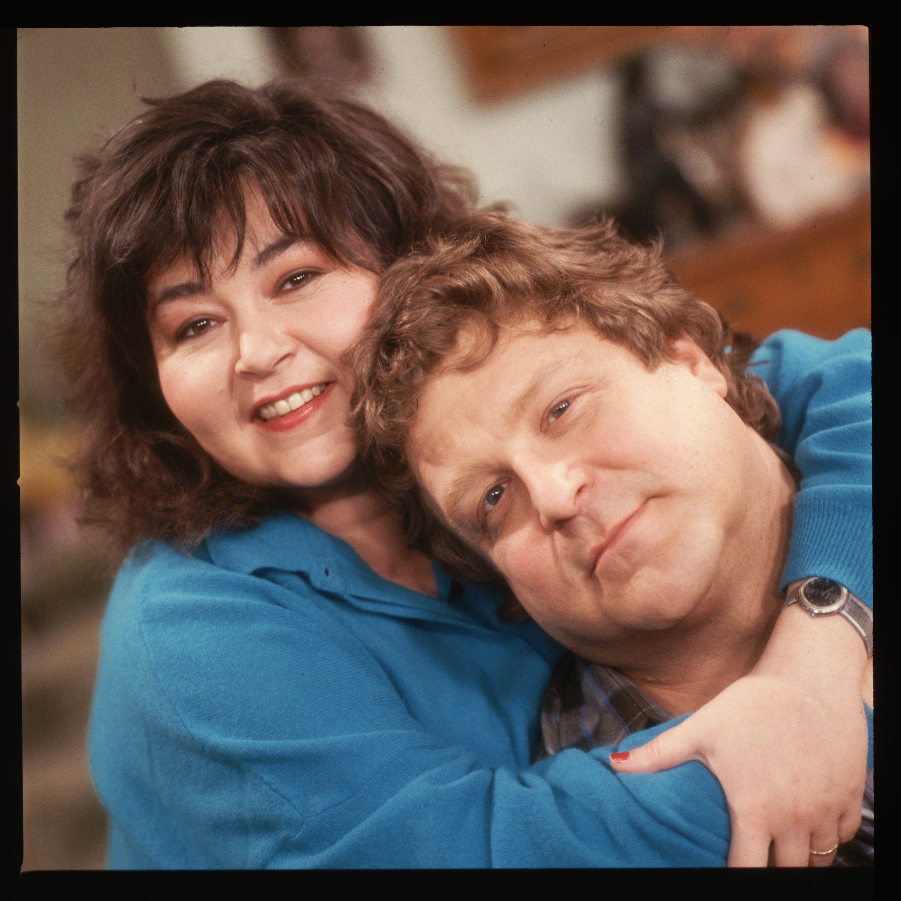Roseanne and John Goodman smiling, 1  hugging the other. Casual clothing, affectionate pose.