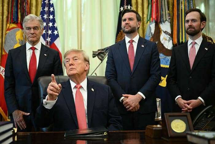 A group of four men in suits stands in an official-looking room, with one seated at a desk, pointing upwards. Flags and medals are displayed behind them
