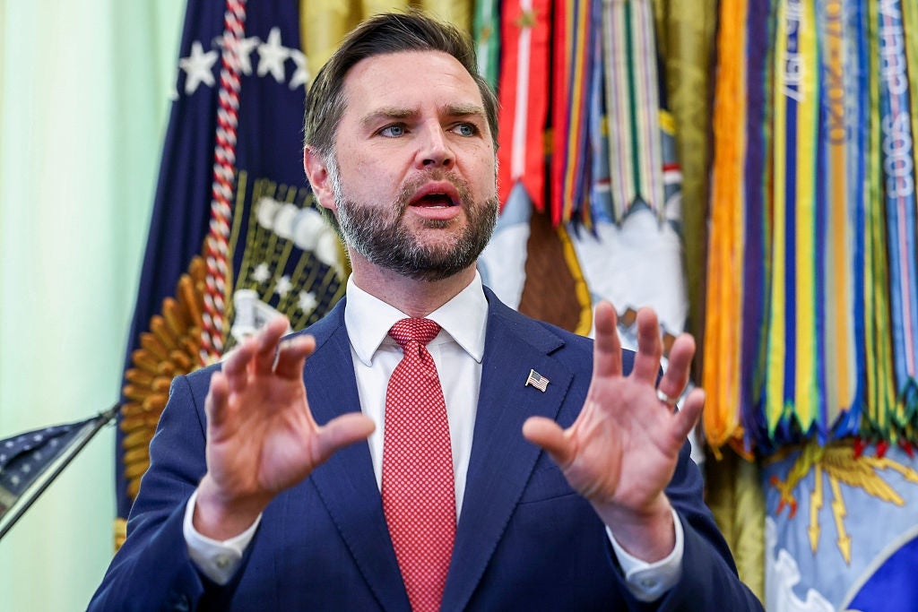 A person in a suit and red tie gestures while speaking, with military flags and emblems in the background