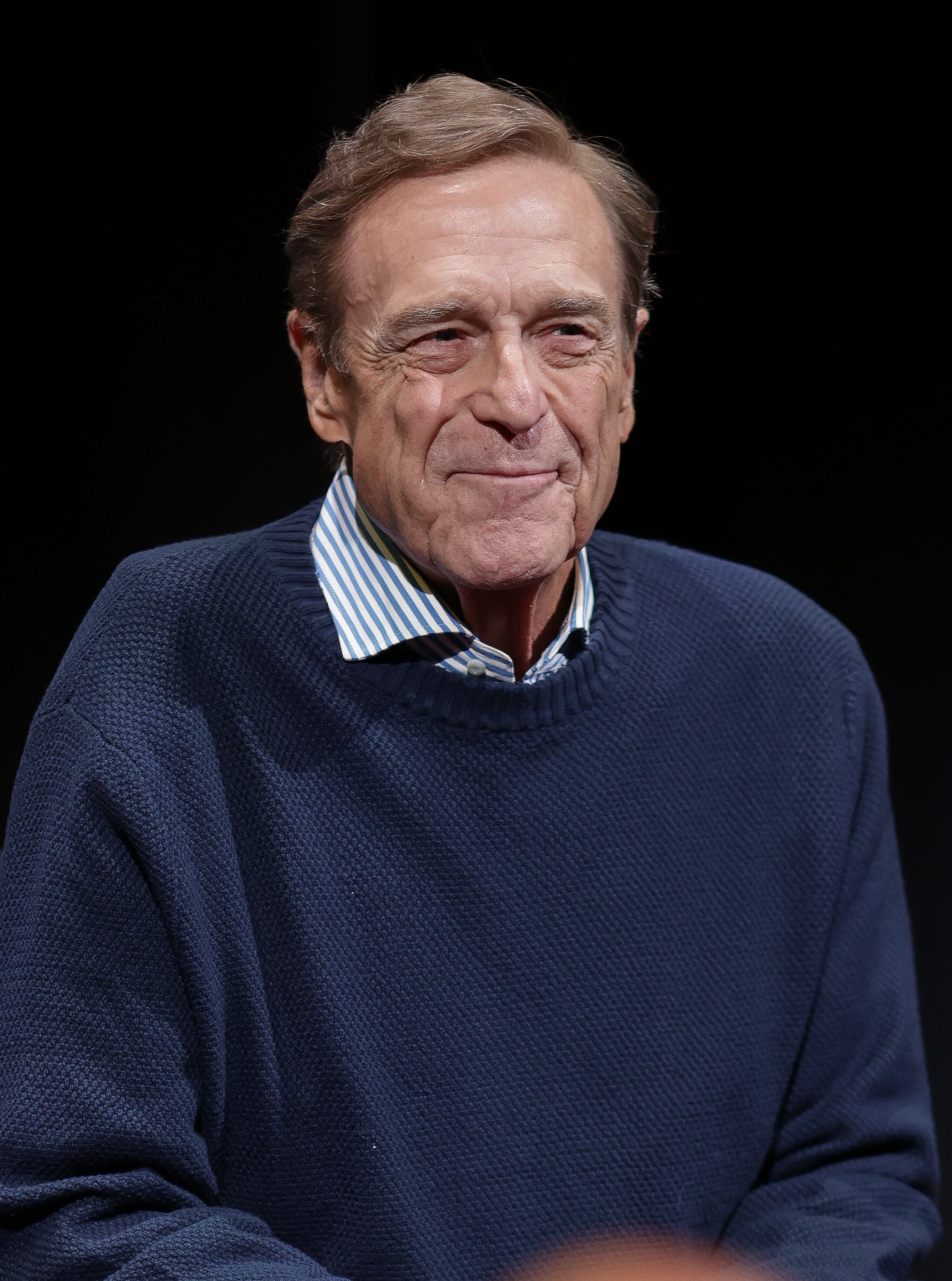 John Goodman wearing a striped garment and acheronian sweater, smiling, seated against a acheronian background