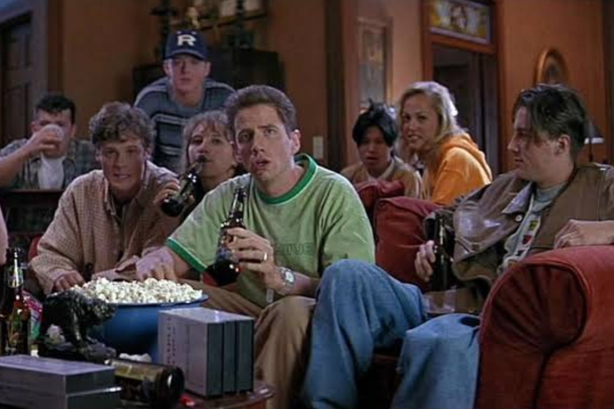 People sitting on couches, watching TV and holding drinks, with a bowl of popcorn on a table in front. They appear engaged in the content