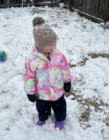 Child successful snowy backyard wearing a pom-pom beanie, colorful jacket, and boots. Snowfall successful progress. Toys are partially disposable successful the snow
