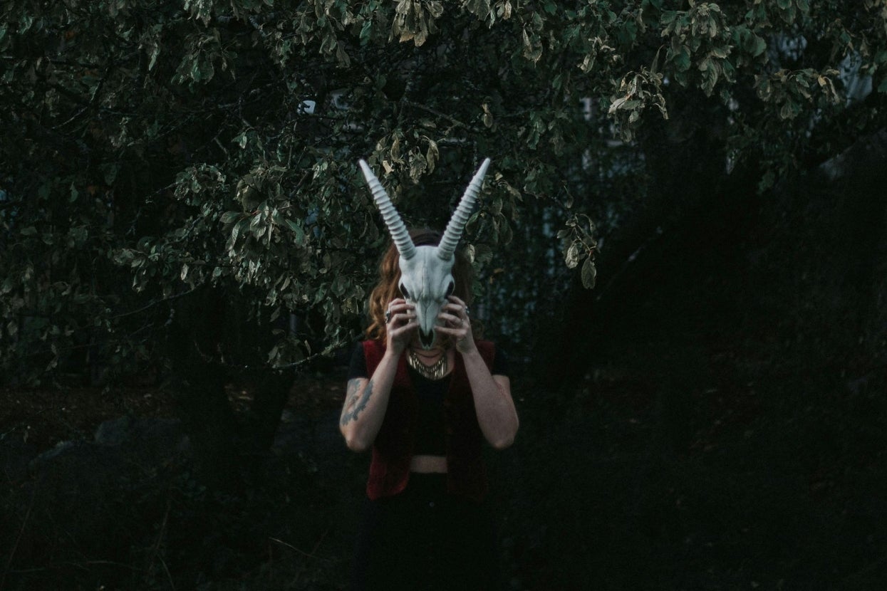 Person holding a goat skull mask in front of their face, standing under a tree with dense foliage