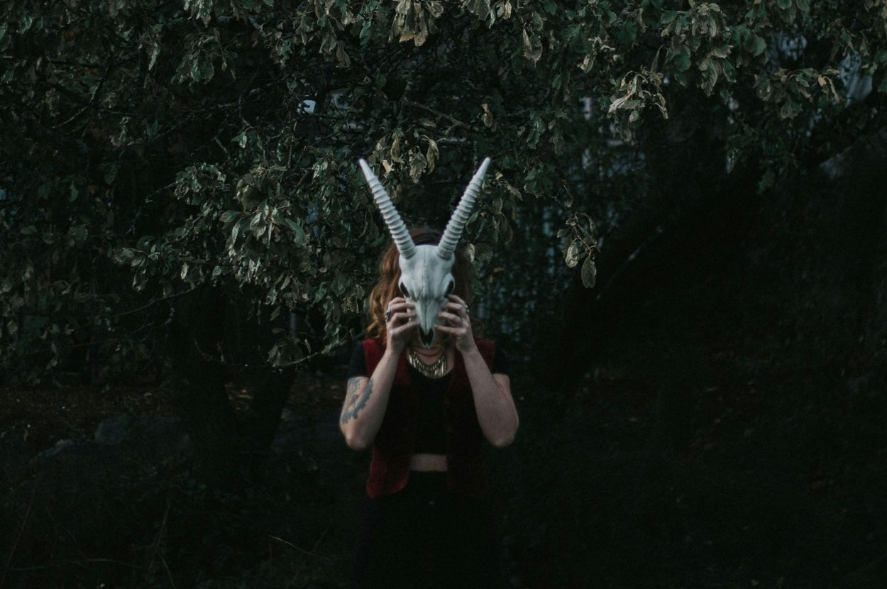 Person holding a goat skull mask in front of their face, standing under a tree with dense foliage