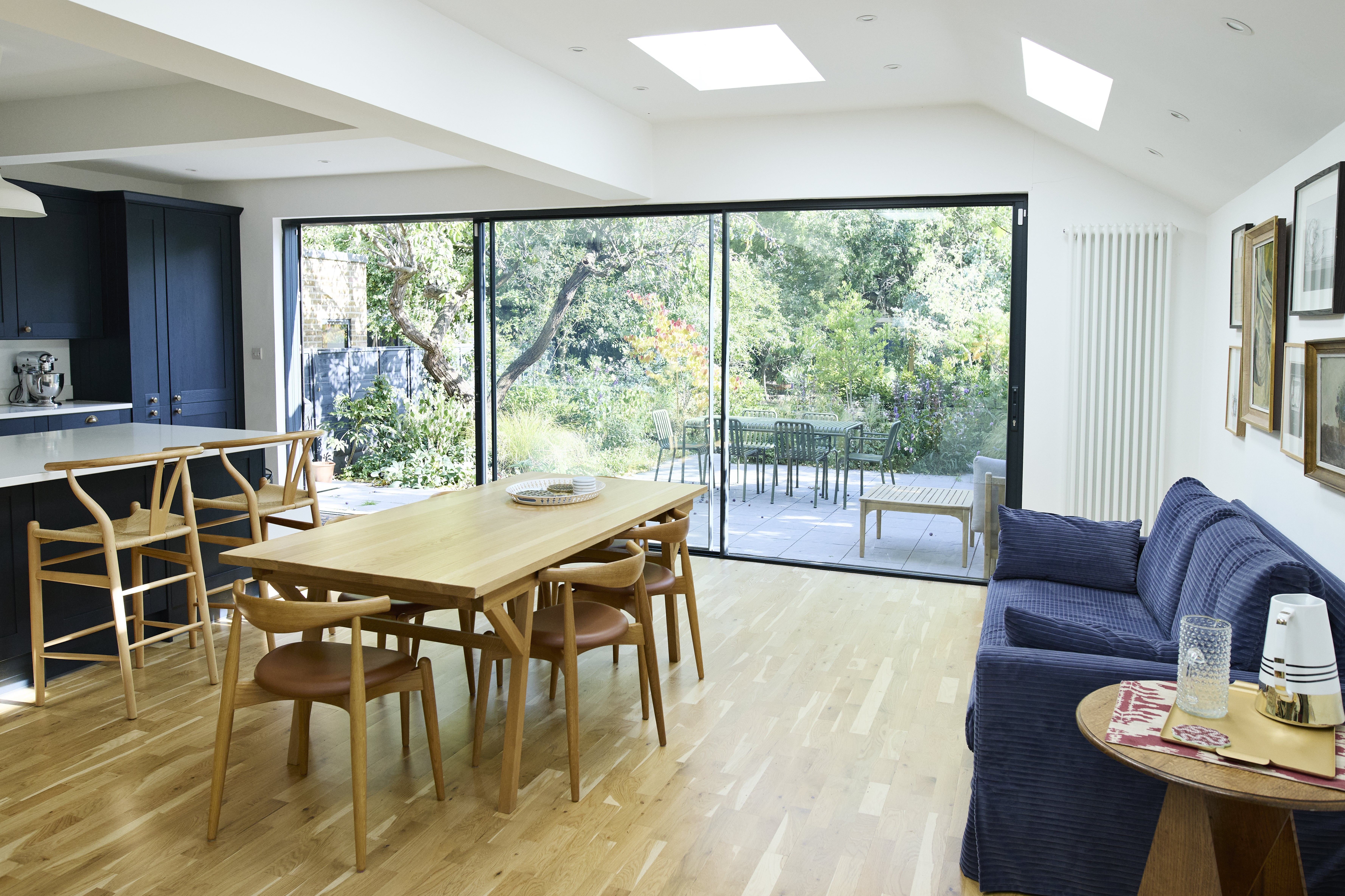 Spacious dining room with wooden furniture, adjacent to a patio with garden view through large glass doors. Cozy blue sofa and modern kitchen area