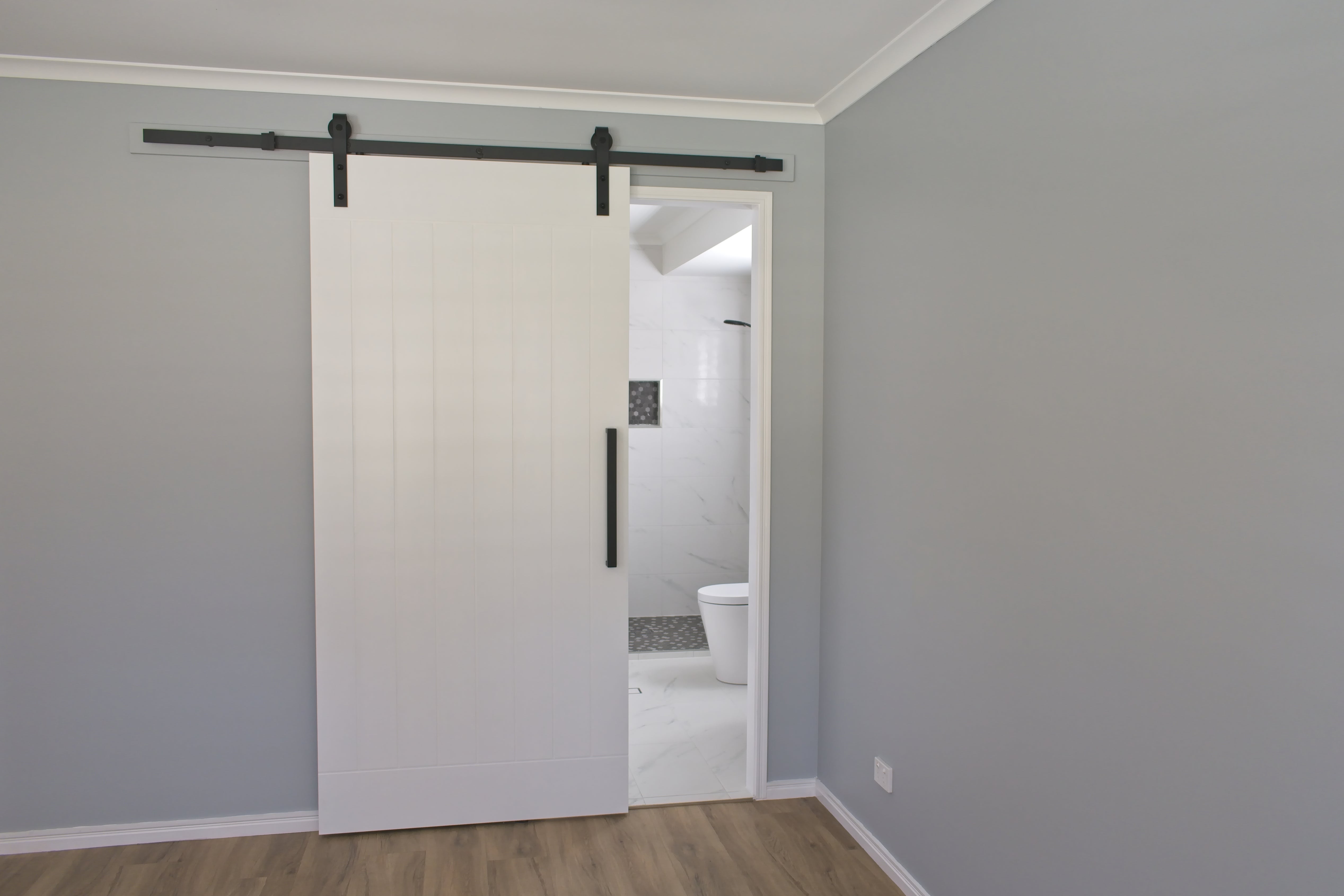 Sliding barn door partially open, revealing a modern bathroom with visible toilet and tiled wall. Room features light wood flooring