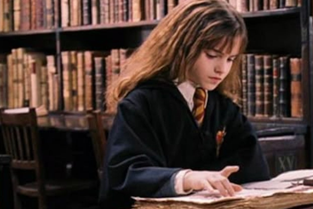 Young Hermione from "Harry Potter" in a school robe and tie reads a large book in a library with shelves of books behind her
