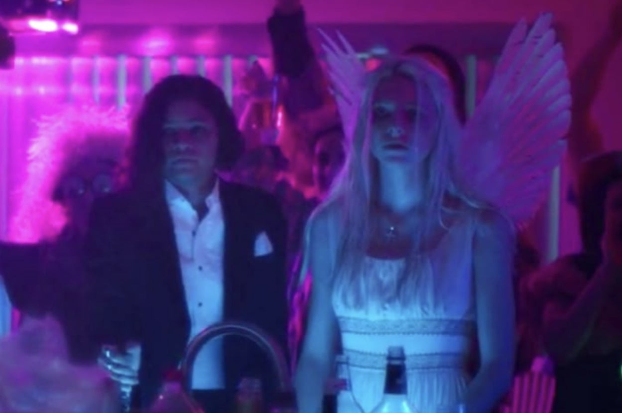 People dressed in party outfits with one wearing angel wings, surrounded by purple lighting in a crowded indoor party setting