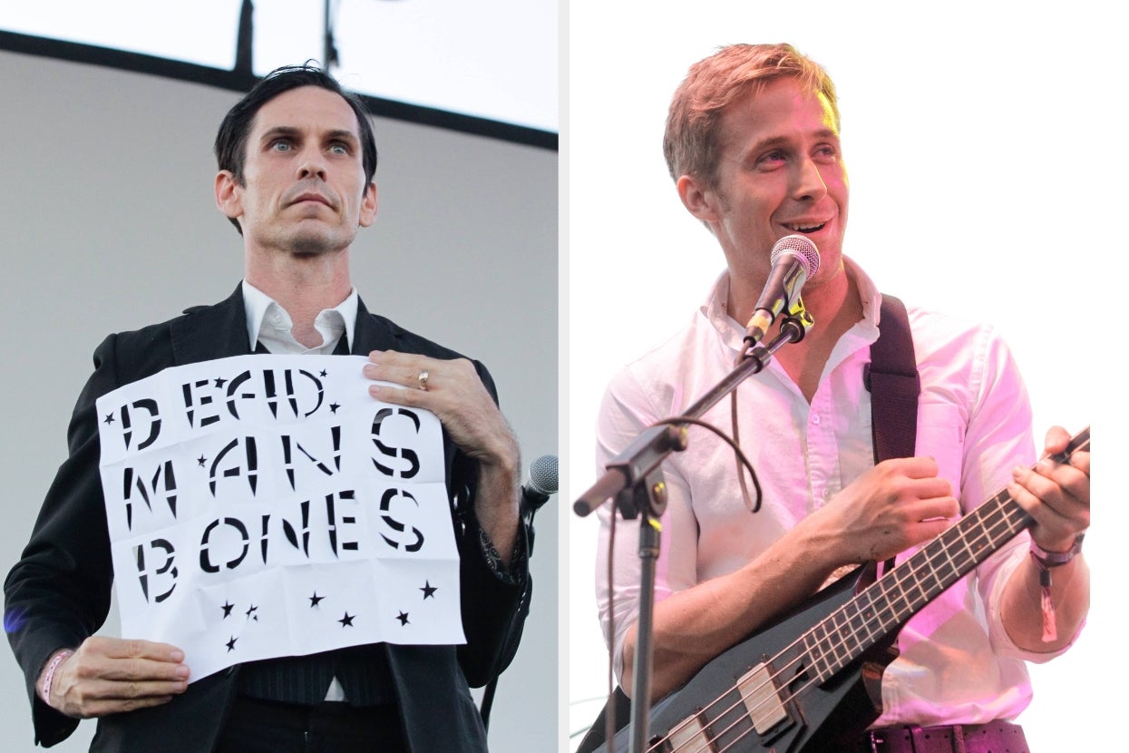 Two performers on stage. The left holds a sign reading "DEAD MAN'S BONES," wearing a black suit. The right plays guitar, wearing a white shirt, smiling