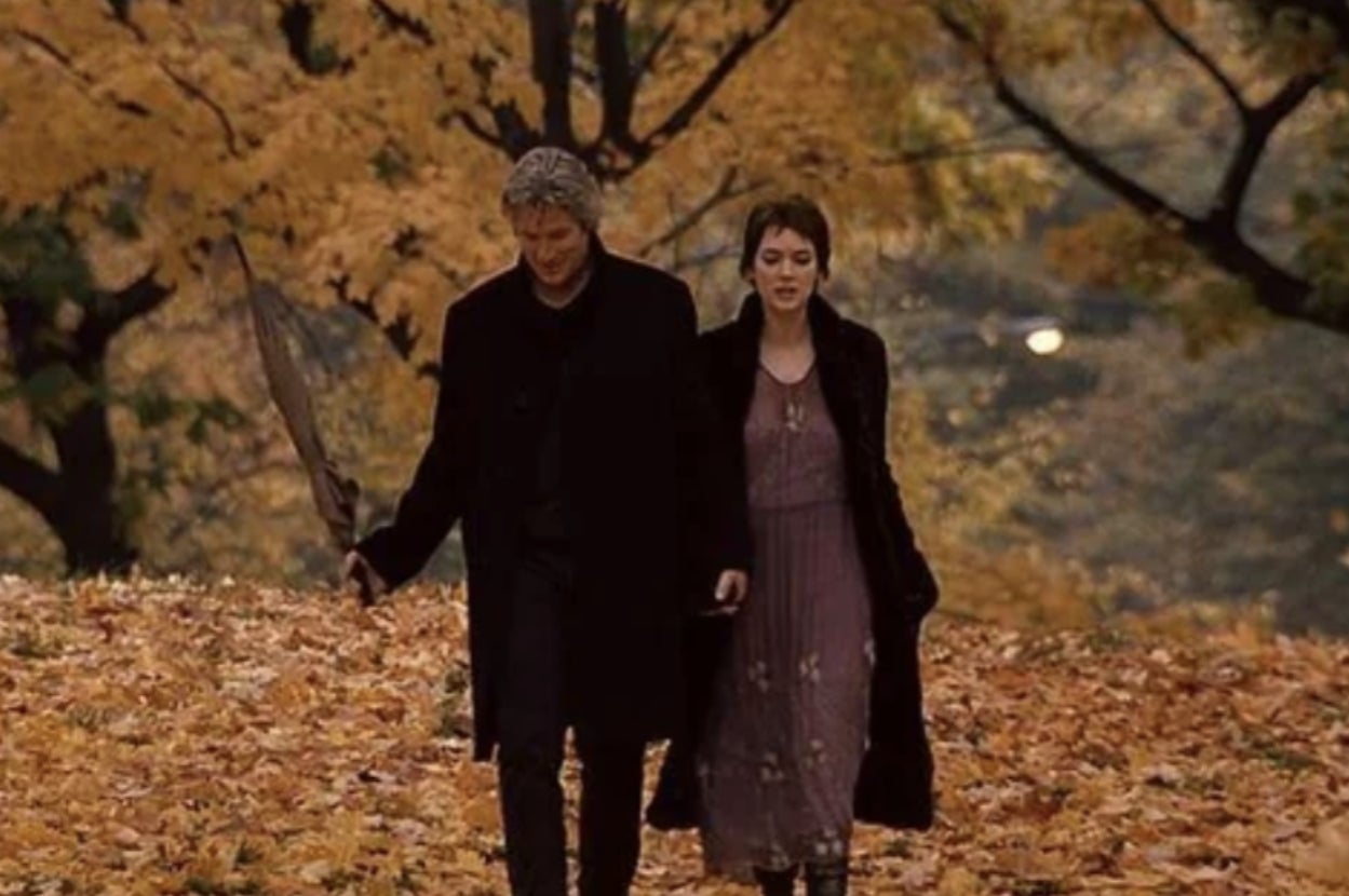A couple strolls through a park with autumn leaves on the ground. The man carries an umbrella, and the woman wears a flowing dress and coat
