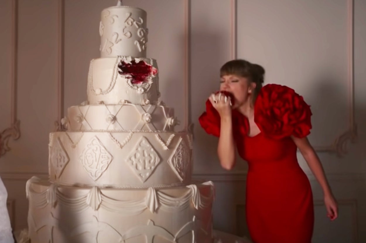 A woman in an elegant red dress playfully takes a bite out of a large, ornate multi-tiered cake