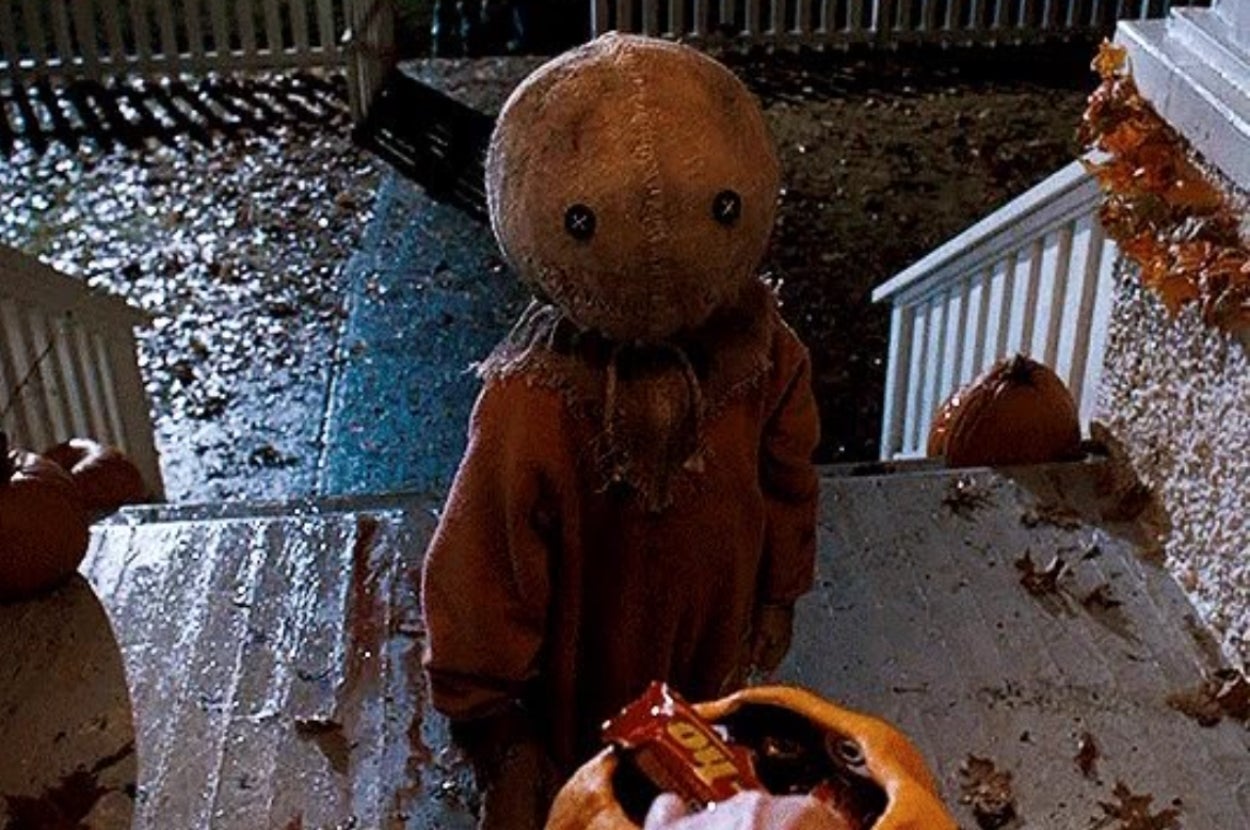 Sam from the movie "Trick 'r Treat" in burlap sack mask stands on porch surrounded by pumpkins, eagerly looking at candy in a trick-or-treat bag