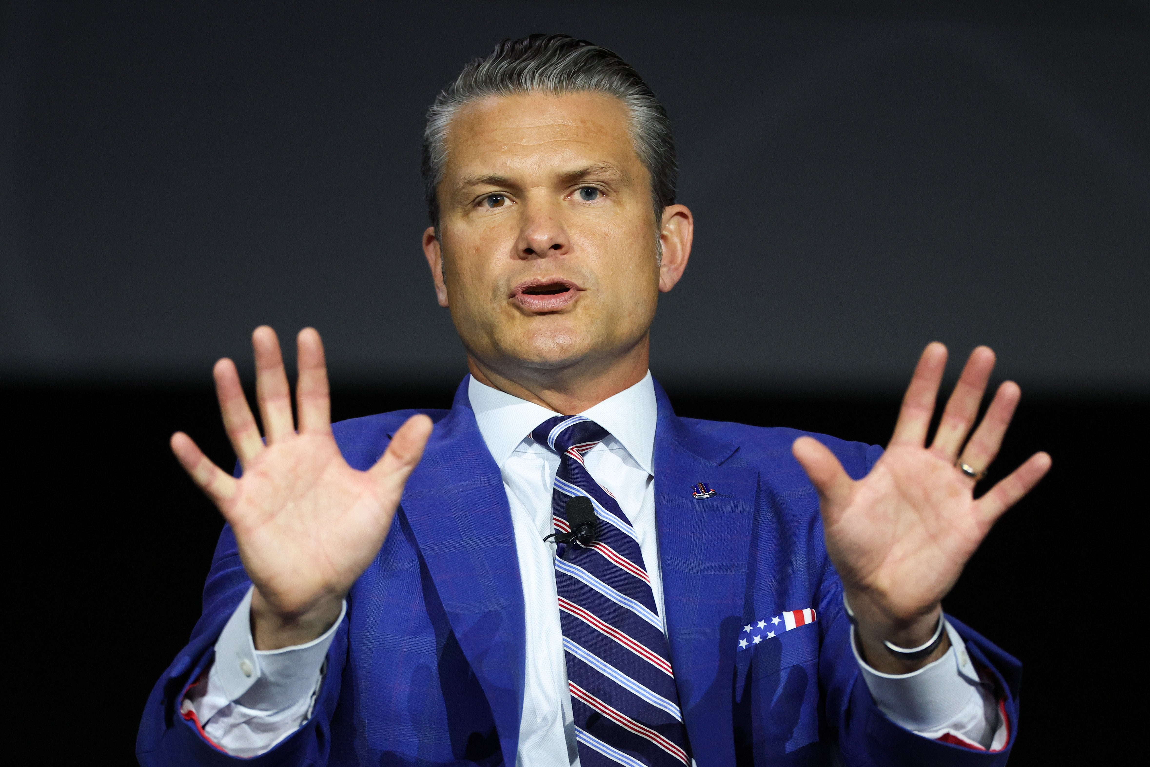 A man in a suit and striped tie is speaking, gesturing with both hands raised in front of him