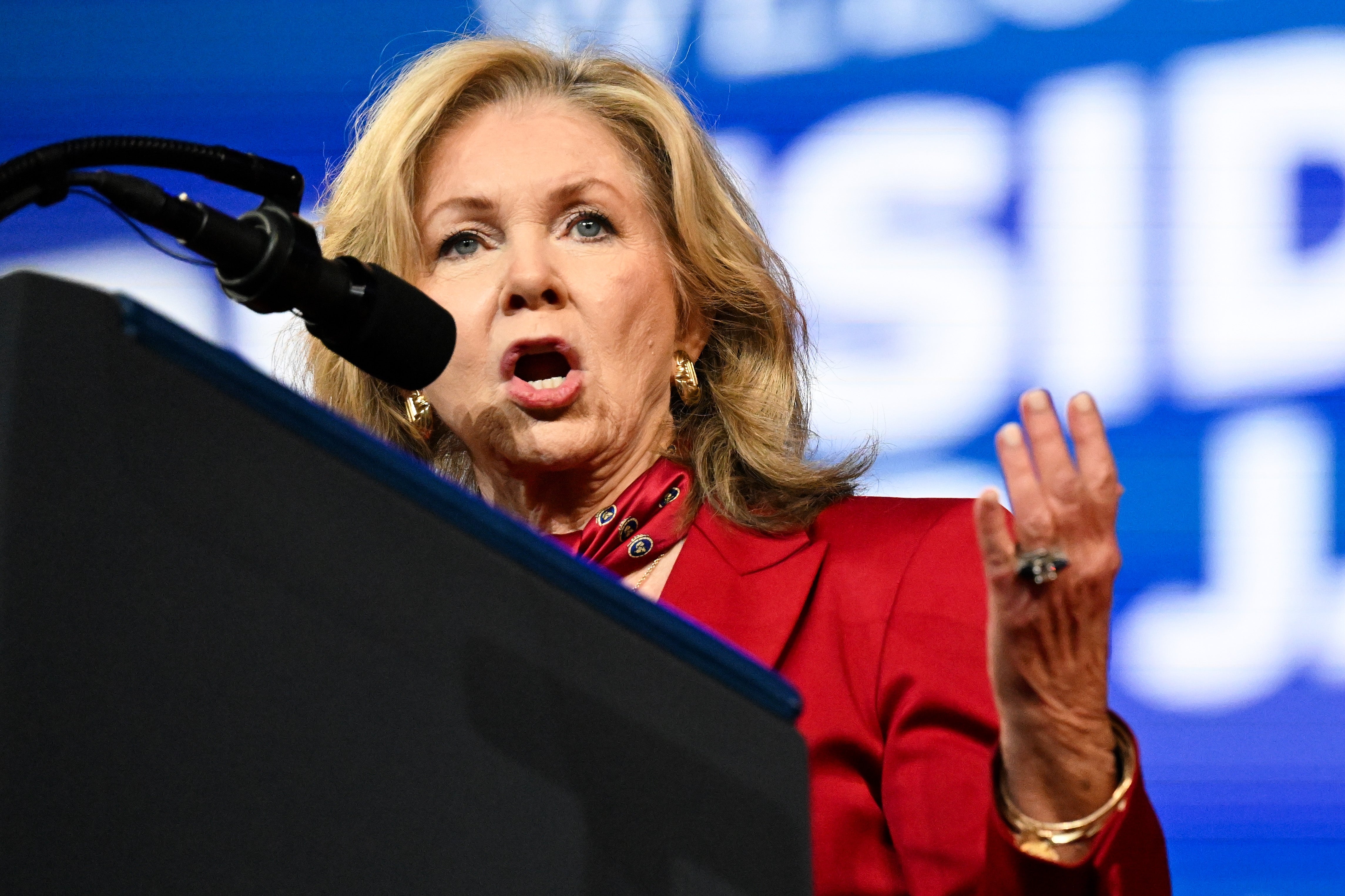 Person speaking passionately at a podium, wearing a red suit and red scarf