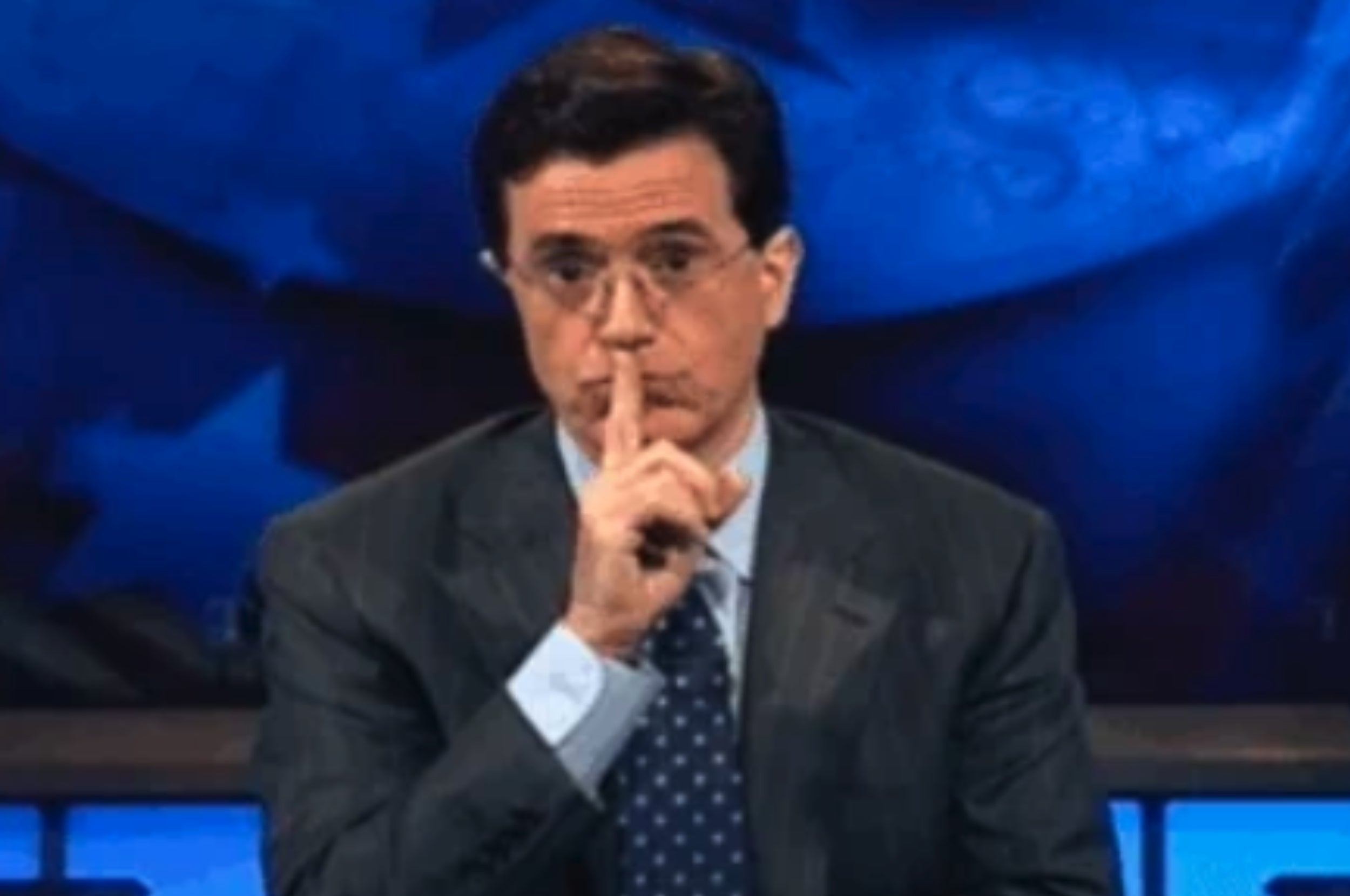 Person in glasses and suit gestures for silence, holding index finger to lips, sitting in a studio setting with a serious expression