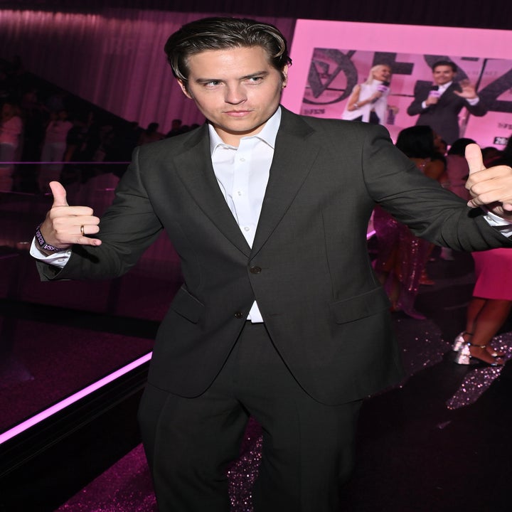 Person in a suit strikes a playful pose with thumbs up at a formal event, in front of a screen displaying people on stage