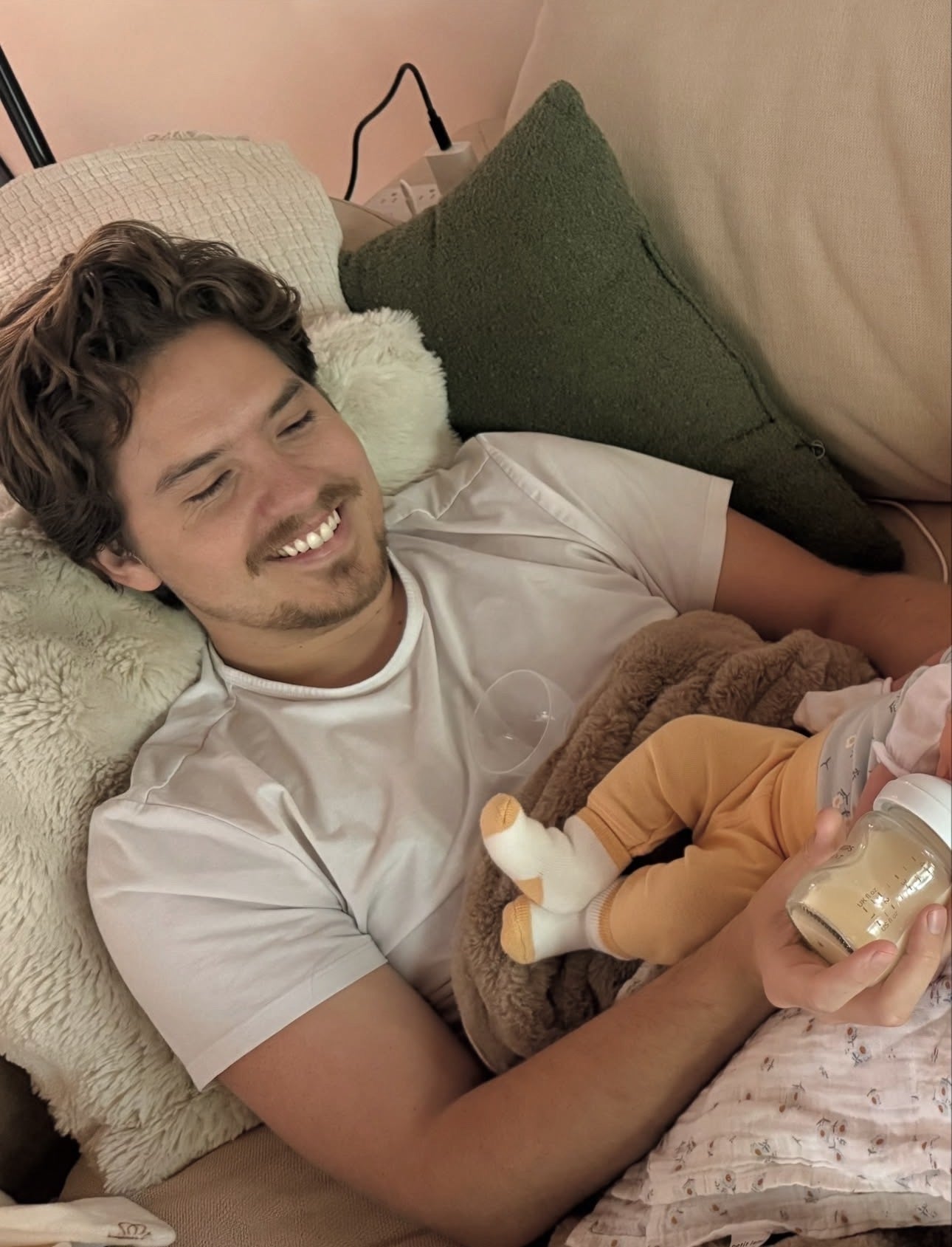 Person lying connected  a sofa  holding a baby, smiling, and offering a bottle. Cozy mounting  with pillows