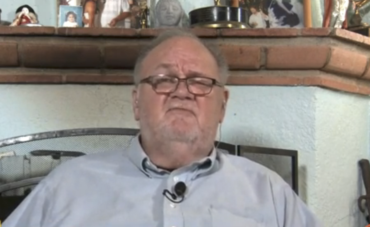Elderly man in a casual shirt sits in front of a shelf with family photos and various items in the background