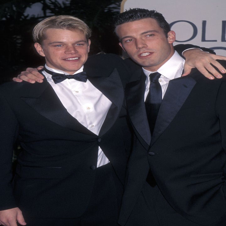 Two men in tuxedos pose together at a formal event, smiling and looking at the camera