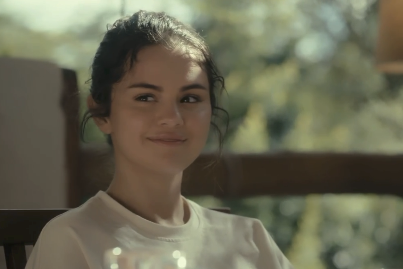 A woman with dark hair smiles gently while sitting outdoors in a relaxed setting