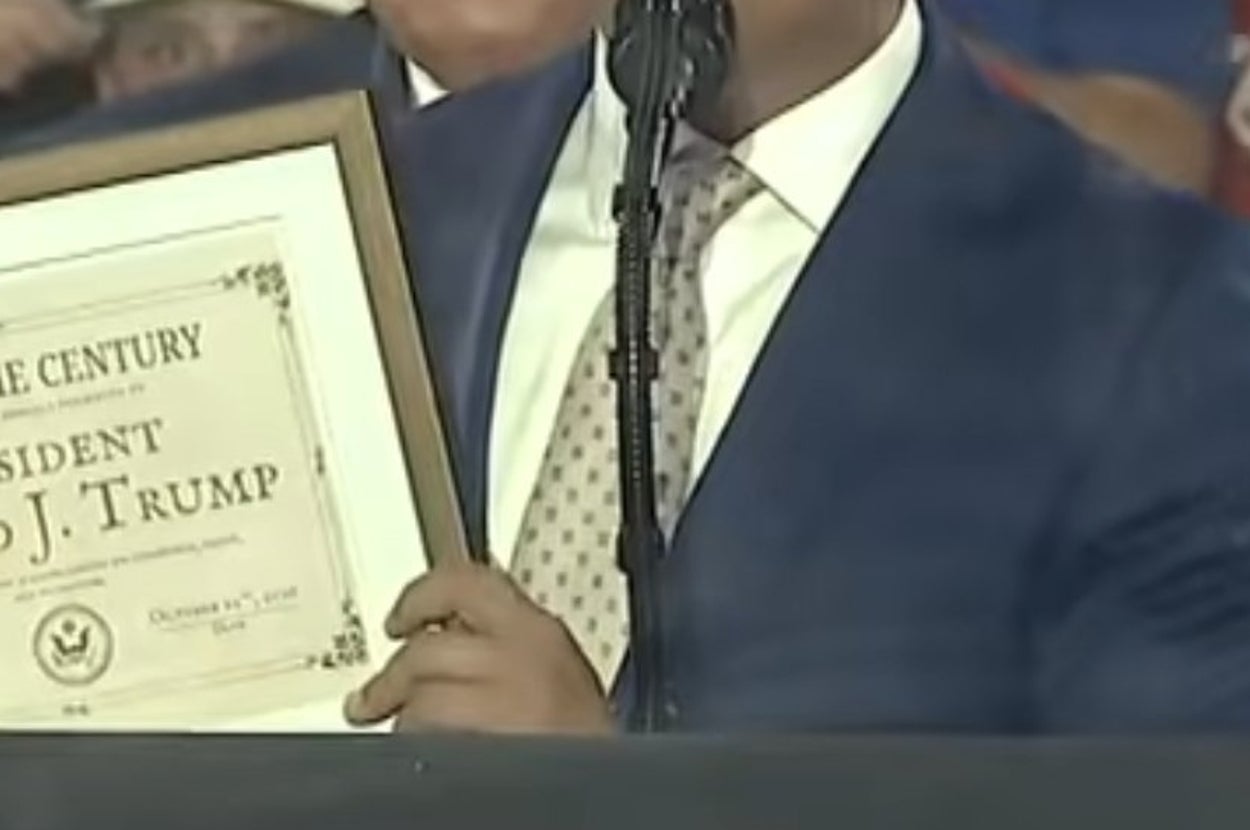 A man in a suit holds a framed certificate labeled "Man of the Century" at a crowded event with people wearing red and holding flags