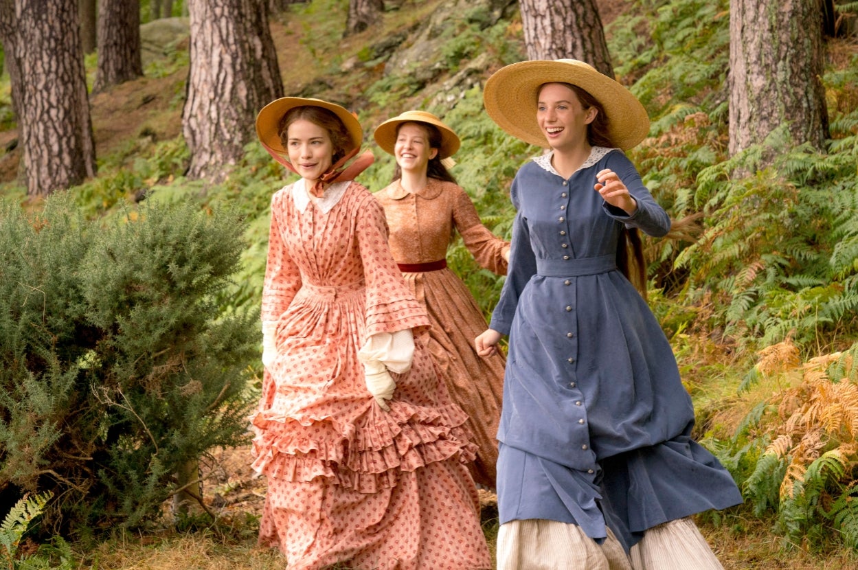 Three people in period costumes and straw hats joyfully walk through a forest setting