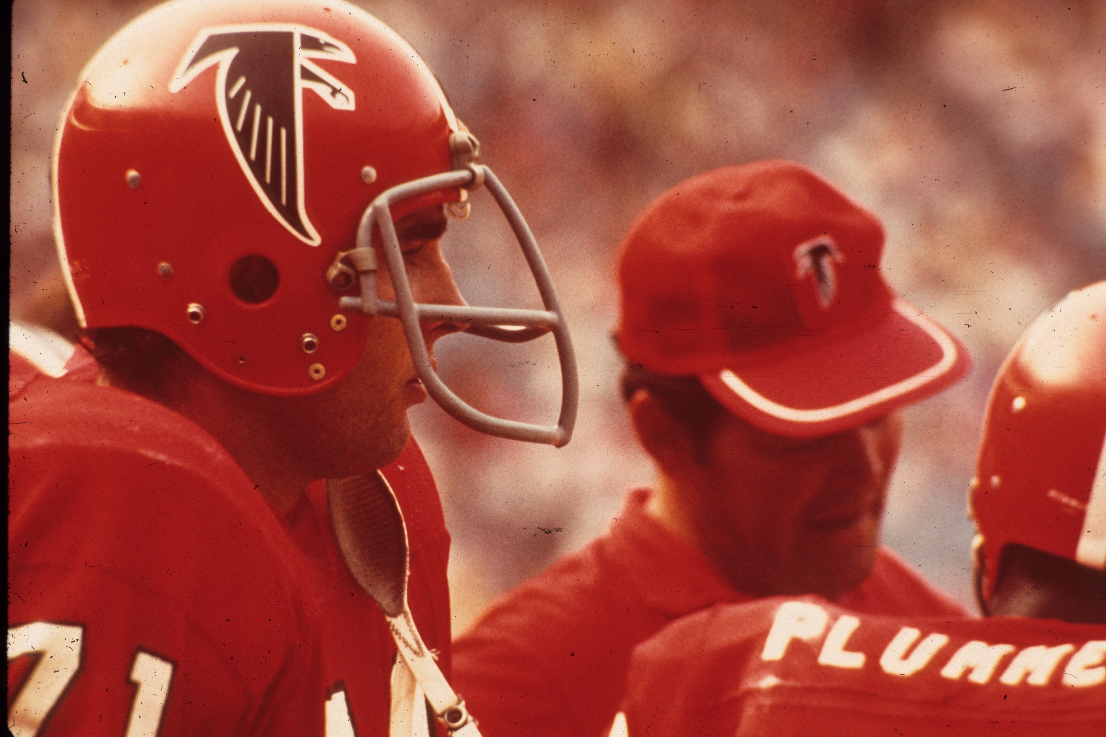 Football players wearing helmets and uniforms are on the sideline. A coach in a cap is speaking to one player