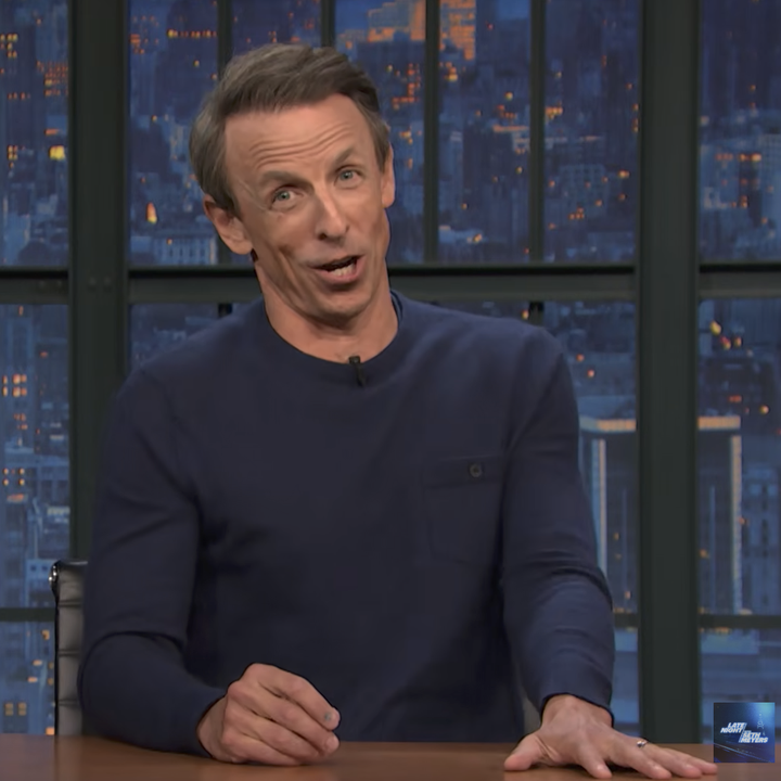 A talk show host sits at a desk gesturing during a nighttime TV broadcast