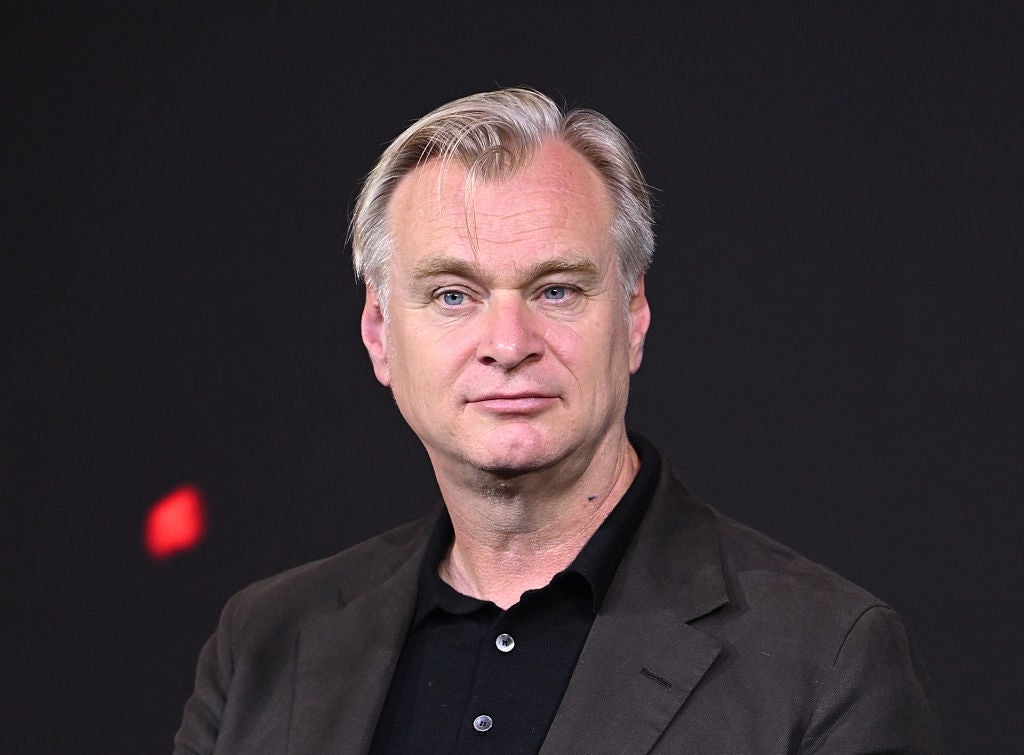 A person is facing the camera, wearing a dark blazer over a button-up shirt, set against a plain background