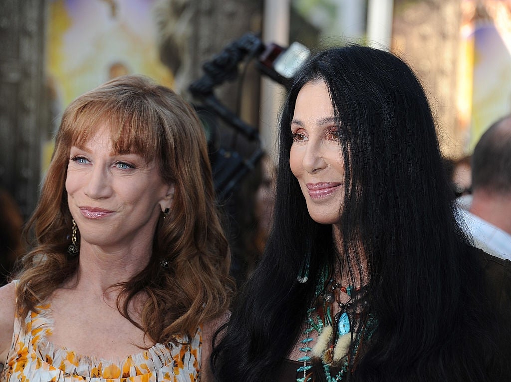 Two people pose on a red carpet. They are wearing stylish outfits, one with patterned details and the other with turquoise jewelry accents