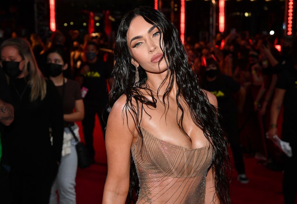 Person in a sheer gown on a red carpet, wet hair styled, surrounded by masked individuals and red lighting