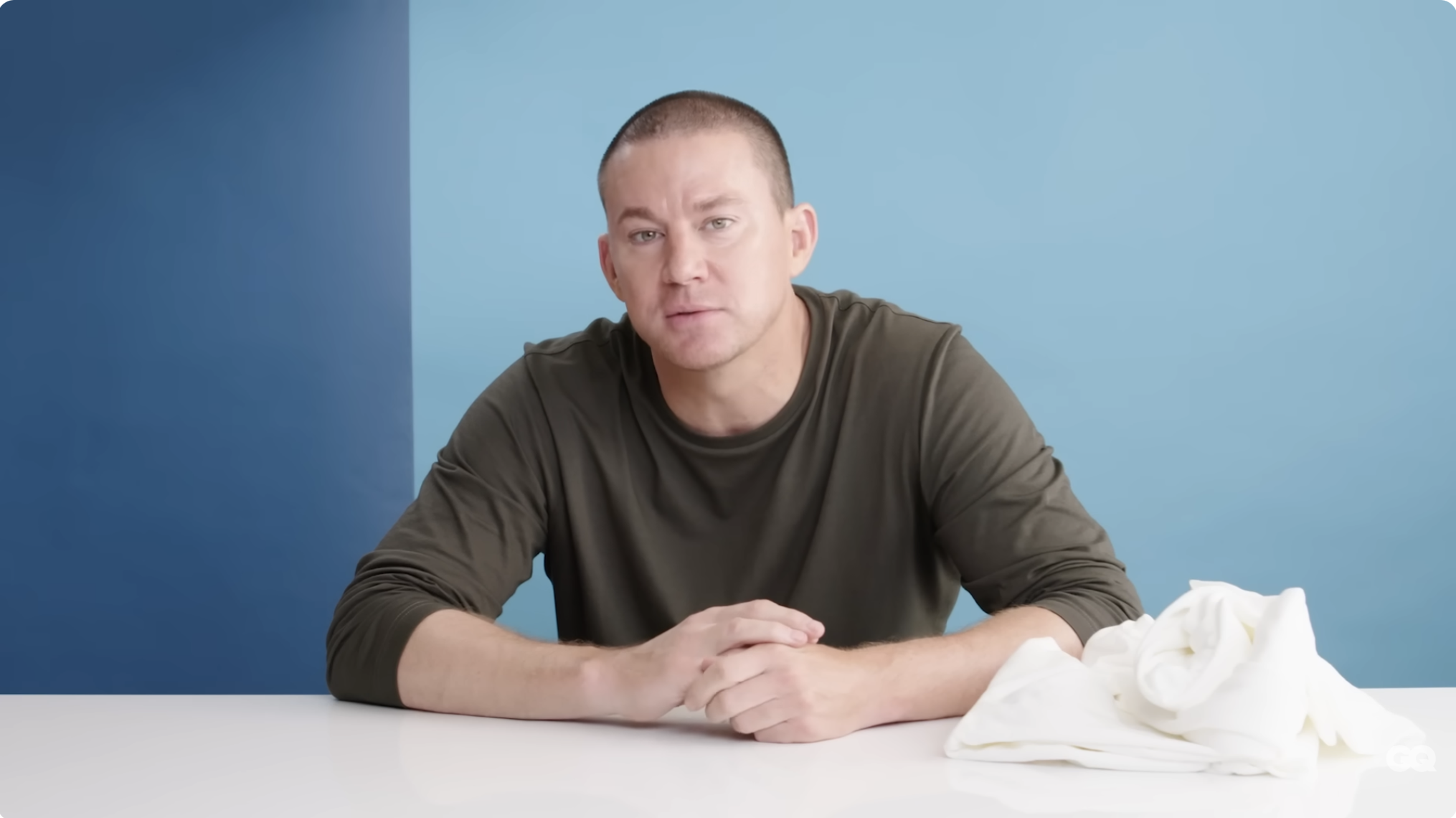 Man seated at a white table against a plain background, wearing a casual, long-sleeved shirt