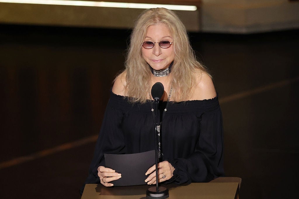 A person with long hair is wearing round sunglasses, an off-the-shoulder dress, and a choker, speaking at a podium