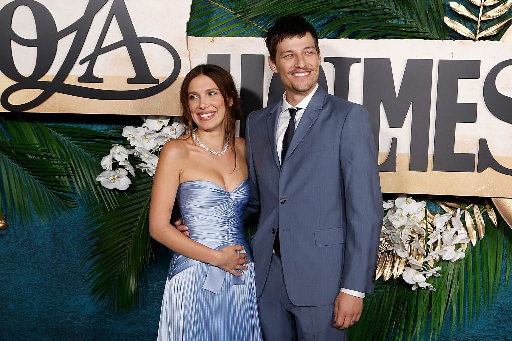Two people pose on a red carpet. The woman wears a strapless gown, and the man wears a suit and tie