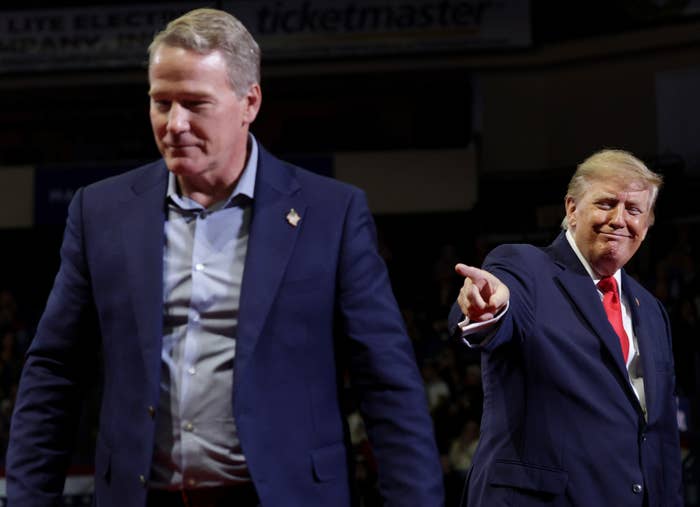 Two individuals astatine  an event; 1  is pointing. Both are successful  suits, 1  with a reddish  necktie  and the different   with an unfastened  garment  collar