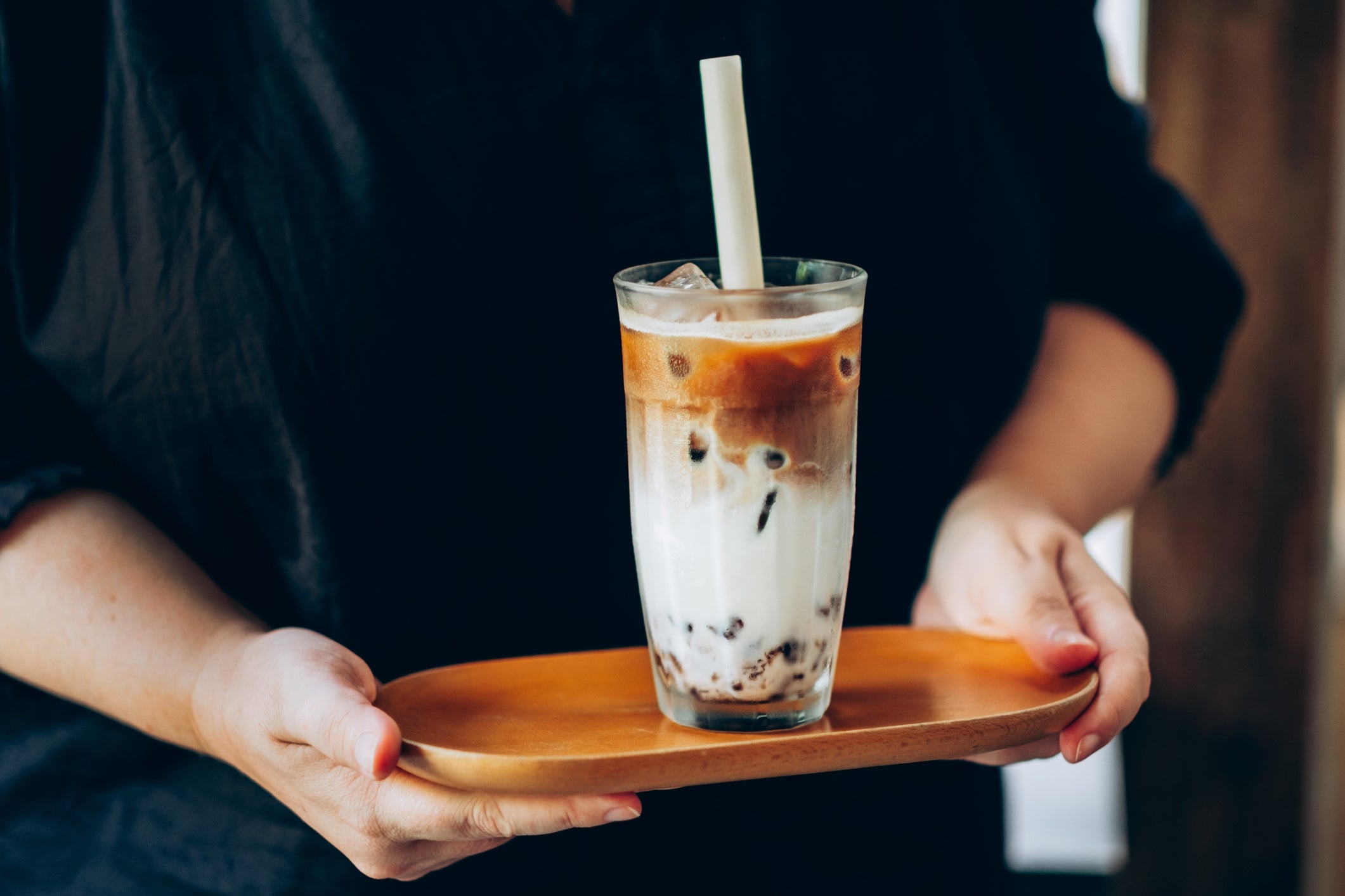 A person holds a wooden tray with a glass of iced coffee topped with milk and tapioca pearls, featuring a large straw