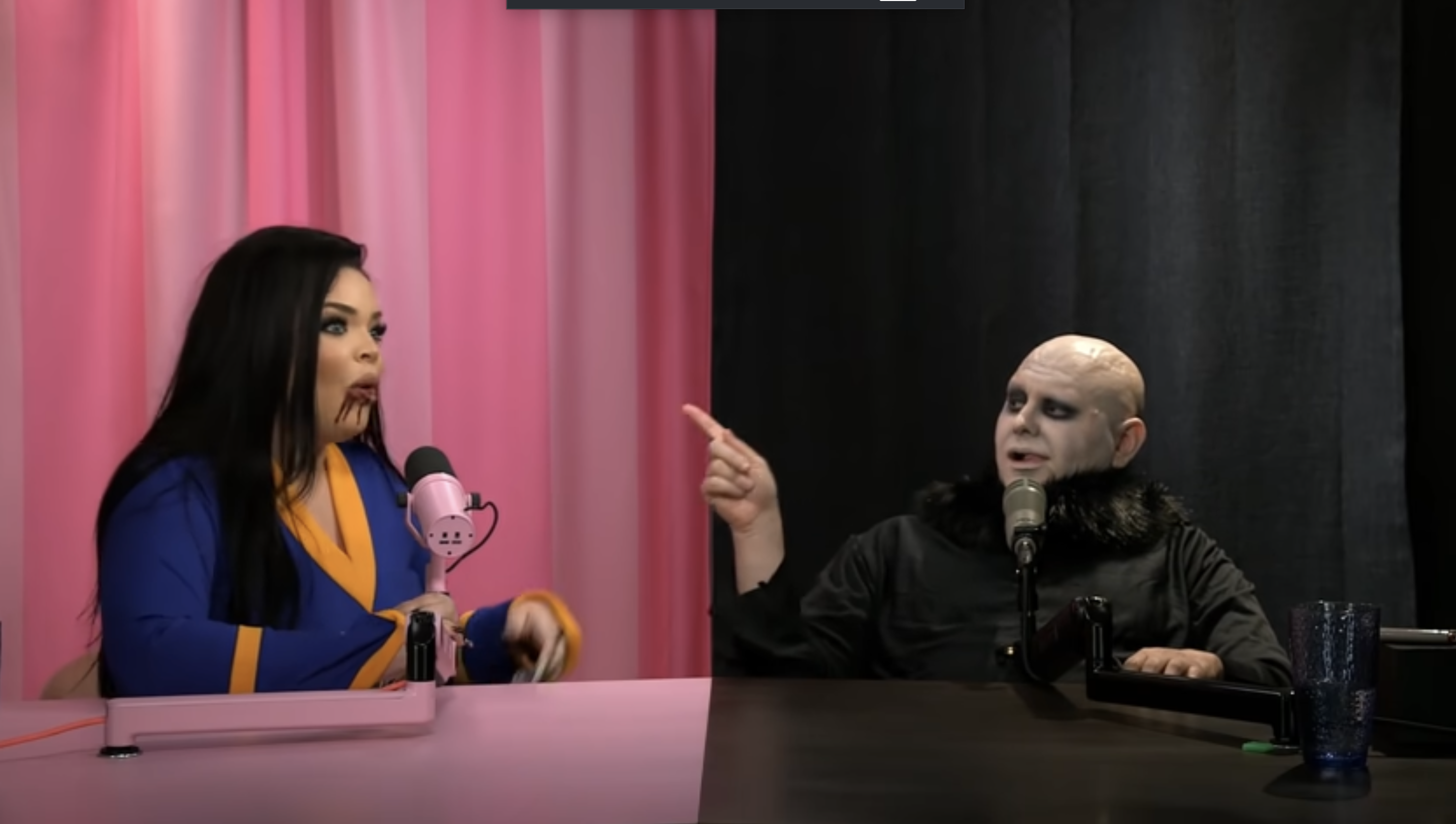Two people in a podcast studio, one with long hair wearing a colorful outfit, and the other with bald head makeup in a dark outfit, in discussion