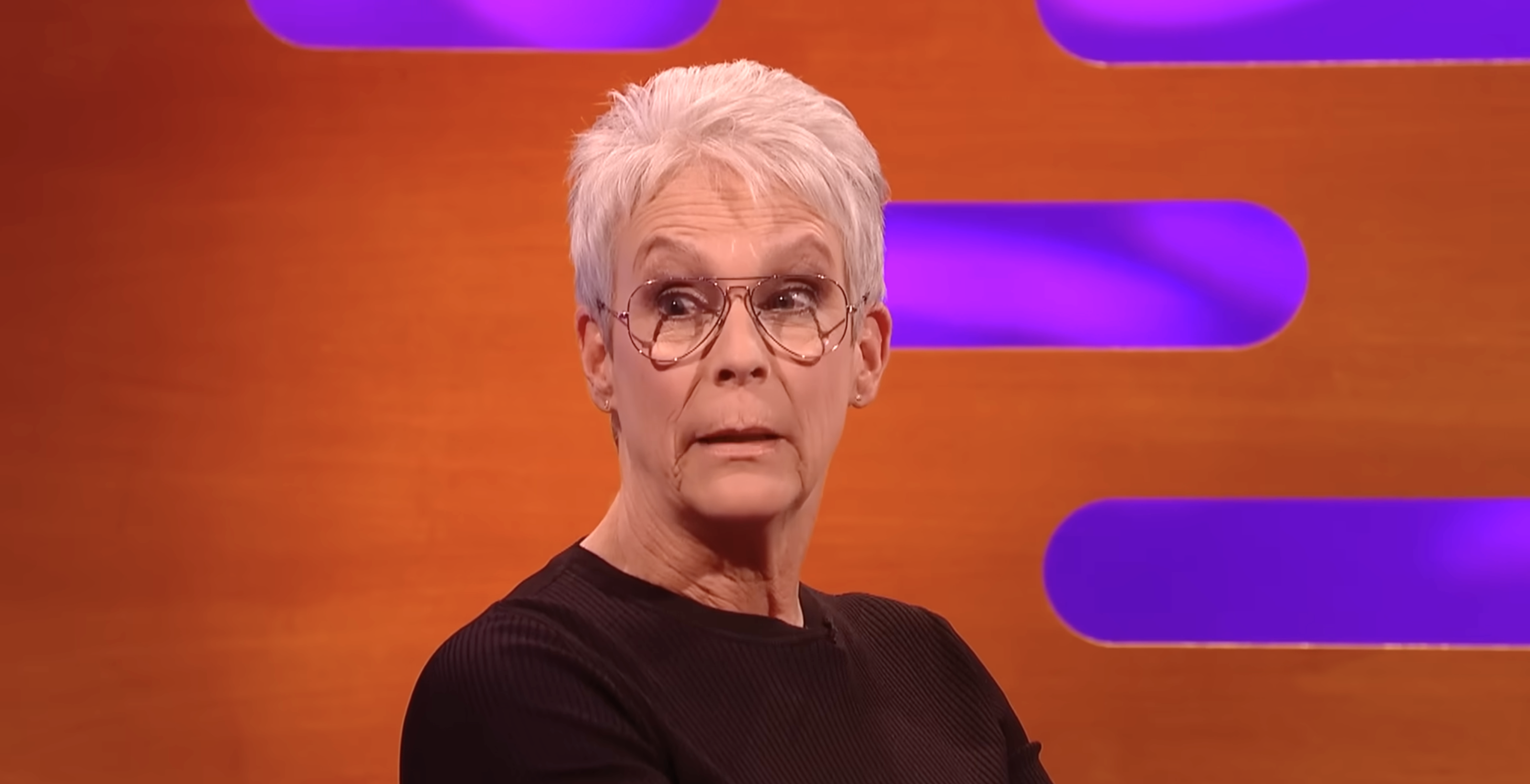 Person with short hair and glasses, wearing a simple black outfit, sitting in front of a backdrop with purple lights, looking sideways