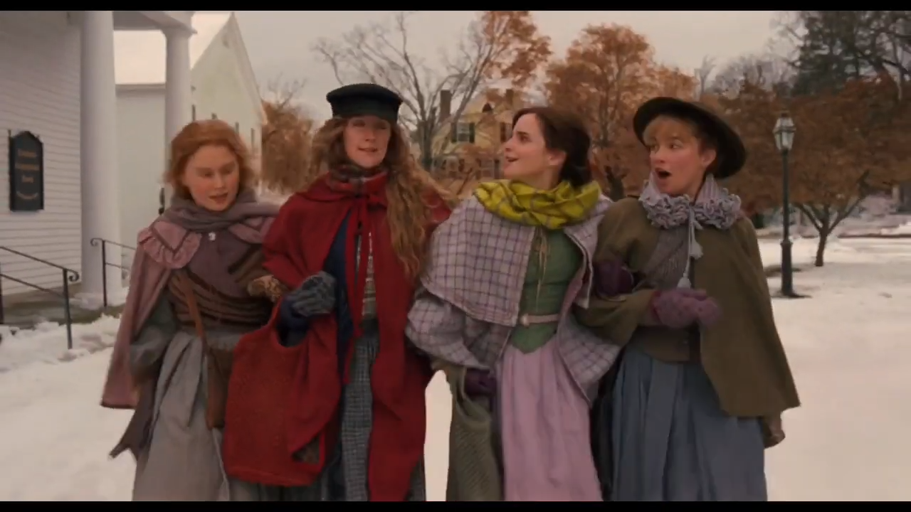 Four women in period costumes walk together outdoors, dressed warmly for winter, with a snowy landscape and bare trees in the background