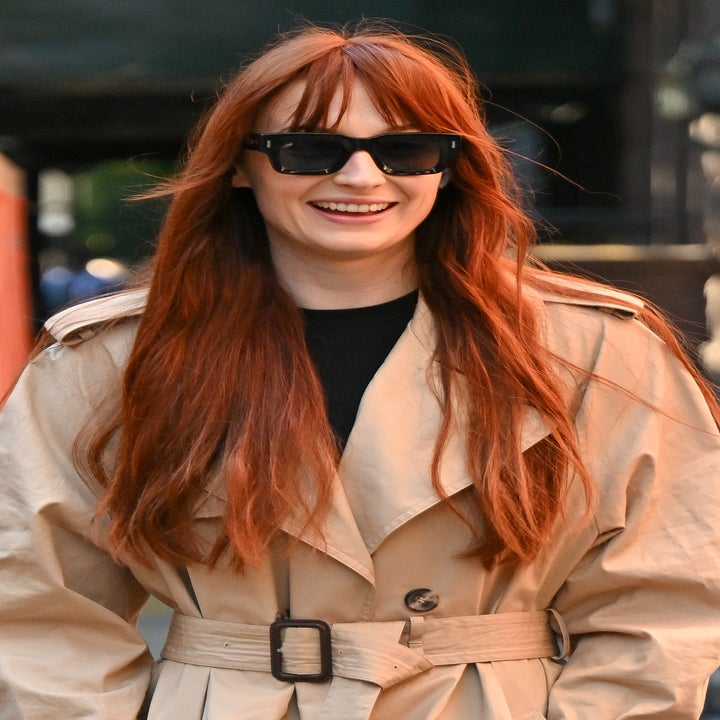 Person with long hair wearing a belted trench coat and sunglasses, smiling outdoors