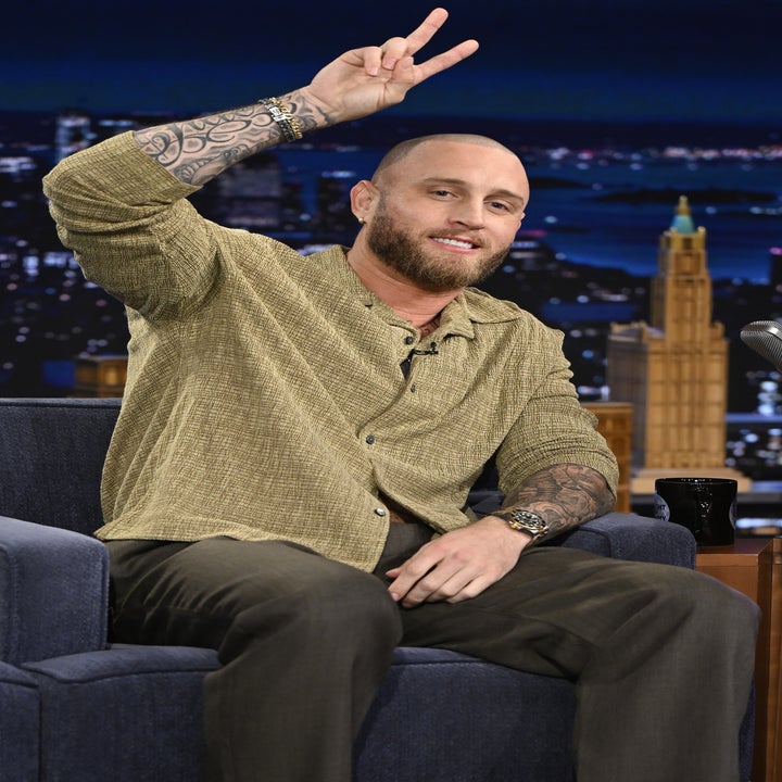 A man with tattoos is seated on a talk show set, wearing a casual button-up shirt and pants, making a peace sign
